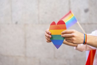 Person's hands holding a rainbow colored heart and a small pride flag, with a rainbow bracelet on