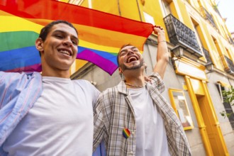 Happy young gay couple holding a rainbow flag and cheering loudly during a pride parade, expressing