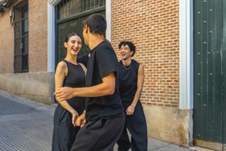 Young friends in black outfits laughing and dancing together on a city street by a brick wall,