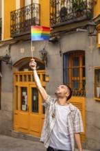 Young man standing in a city street, raising a small rainbow flag with arm outstretched,