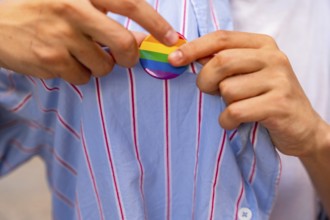 Person's hands carefully pinning a rainbow pride flag button onto a blue and white striped shirt,