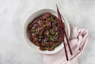 Crispy beef, with green onions and sesame seeds, in a bowl with chopsticks, on a textured surface,