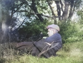 Colourised image of pensive young man lying in countryside field with trees c 1910-1920
