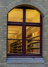 A view through the window of a library, shelves full of books illuminated by a warm, yellow light,