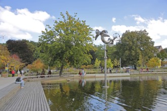 Bastinsweiher with sculpture, seating, bench, people, pedestrians, park, pond, autumn colours,