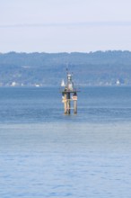 A lighthouse in blue water with a sailing ship, green hills and clear sky in the background,