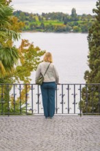 Person standing at the railing of a walkway and looking at a lake surrounded by trees in autumn,