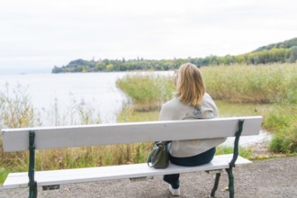 Woman on park bench looking at calm lake and autumn landscape, Mainau Island, Lake Constance,