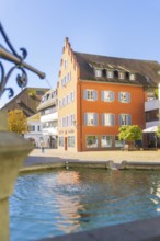 A fountain in the foreground with a striking historical building in the sunlight, Überlingen, Lake