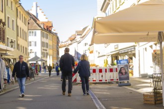 People walk along a sunny street, cafés and shops surround them, Überlingen, Lake Constance,