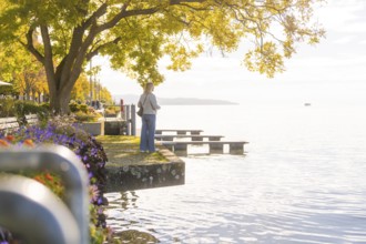 A woman stands on the lakeshore, surrounded by trees and flowers, while the scenery is bathed in
