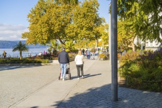 People walking on a sunny promenade along autumnal trees by the water, Überlingen, Lake Constance,