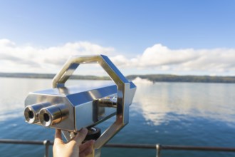 Hand holding binoculars on the lakeshore under a clear sky with clouds, Überlingen, Lake Constance,