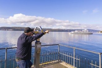Man pointing with binoculars at a ship on a calm lake under a clear sky, Überlingen, Lake