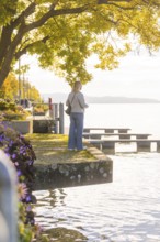 A woman stands on the shore of a lake in a relaxed atmosphere, surrounded by trees and flowers in
