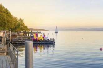 A river with colourful figures and a sailing boat on the calm lake at sunrise, Überlingen, Lake