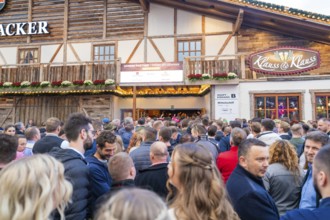 A large crowd waits outside in front of a festively decorated wooden house, Cannstatter Wasen fair,