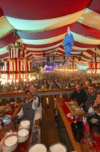 People celebrating at a long table in a colourful striped marquee, Cannstatter Wasen folk festival,