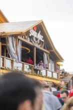 People standing on a balcony of a Bavarian wooden building with a glass front, Cannstatter Wasen