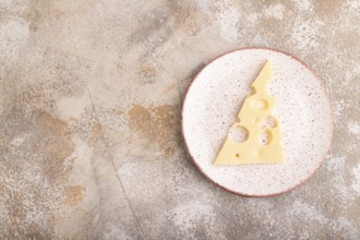 Piece of cheese with holes on ceramic plate on brown concrete background, top view, flat lay, copy