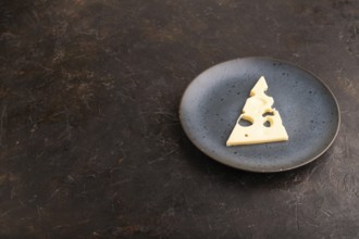 Piece of cheese with holes on blue plate on black concrete background, side view, copy space,