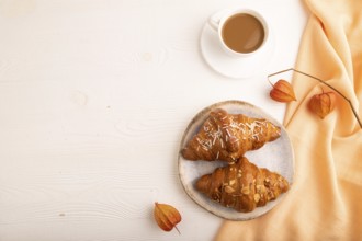 Croissant on blue plate on white wooden background and orange linen textile, cup of coffee, top