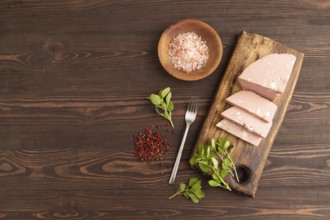 Mortadella sausage on wooden cutting board with pepper and herbs on brown wooden background. Top