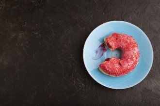 Bitten Pink Donut with sprinkles on blue ceramic plate on black concrete background, top view, flat