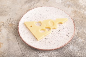 Piece of cheese with holes on ceramic plate on brown concrete background, side view, close up,