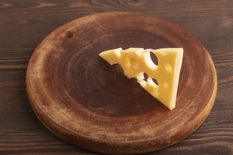 Piece of cheese with holes on cutting board on brown wooden background, side view, close up,