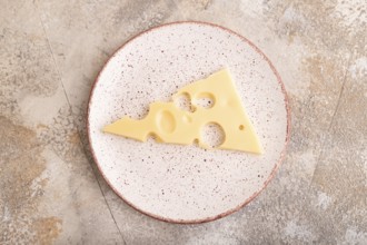 Piece of cheese with holes on ceramic plate on brown concrete background, top view, flat lay, copy