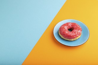 Pink Donut with sprinkles on blue ceramic plate on blue and orange pastel paper background, side