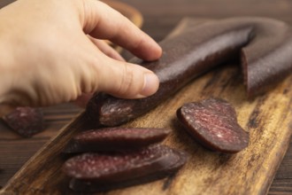 Sujuk sausage with hand on wooden cutting board with pepper and herbs on brown wooden background.
