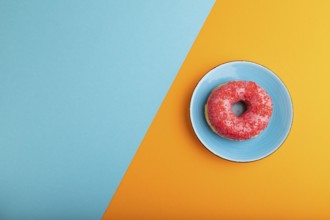 Pink Donut with sprinkles on blue ceramic plate on blue and orange pastel paper background, top