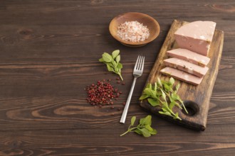 Mortadella sausage on wooden cutting board with pepper and herbs on brown wooden background. Side