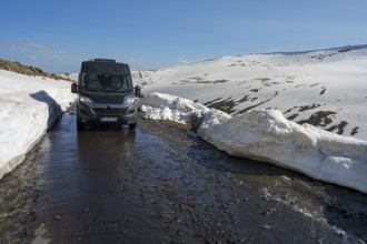 Van auf schmelzender Straße zwischen hohen Schneewänden in bergiger Umgebung, mit dem Camper zum