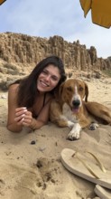 Young woman and her furry companion smiling, lying on sandy beach under an umbrella, enjoying a