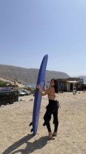 Young woman smiling, holding a blue surfboard on a dusty beach, wearing a wetsuit top and bottom,