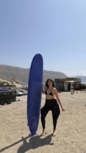 Young woman on a sunny moroccan beach holding a blue surfboard, wearing a bikini top and wetsuit