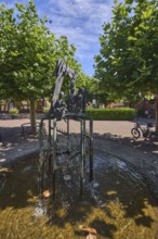 Brunnen mit Metallskulpturen, Alltägliches Theater, Bildhauer Hugo Uhl, Platz, Bäume, Hecke, blauer