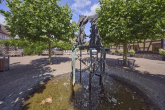 Brunnen mit Metallskulpturen, Alltägliches Theater, Bildhauer Hugo Uhl, Platz, Bäume, Hecke,