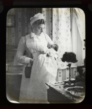 Magic lantern slide of nurse in uniform holding newborn baby inside a home, England, UK c 1900-1910