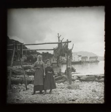 Magic lantern slide of mother and daughter standing by wooden frame of cod fish drying with village