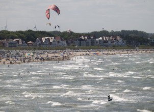 Kitesurfers and holidaymakers on the beach and in the sea, waves, beach chairs, houses in the
