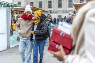 Happy multi ethnic couple walking arm in arm between christmas market stalls, woman carrying