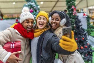 Three cheerful multi ethnic friends taking a selfie at a christmas market while holding a gift,