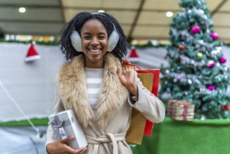 Young black woman wearing earmuffs holding christmas present and shopping bags, smiling in front of