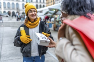 Two friends joyfully exchanging gifts at a bustling christmas market in the city, embracing the