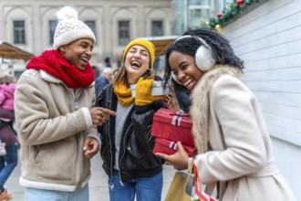 Three friends are having fun exchanging christmas gifts at a winter market, laughing and enjoying