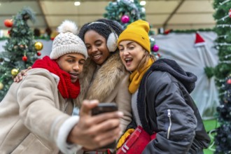 Three cheerful young friends taking a selfie with smartphone in a christmas decorated shopping mall